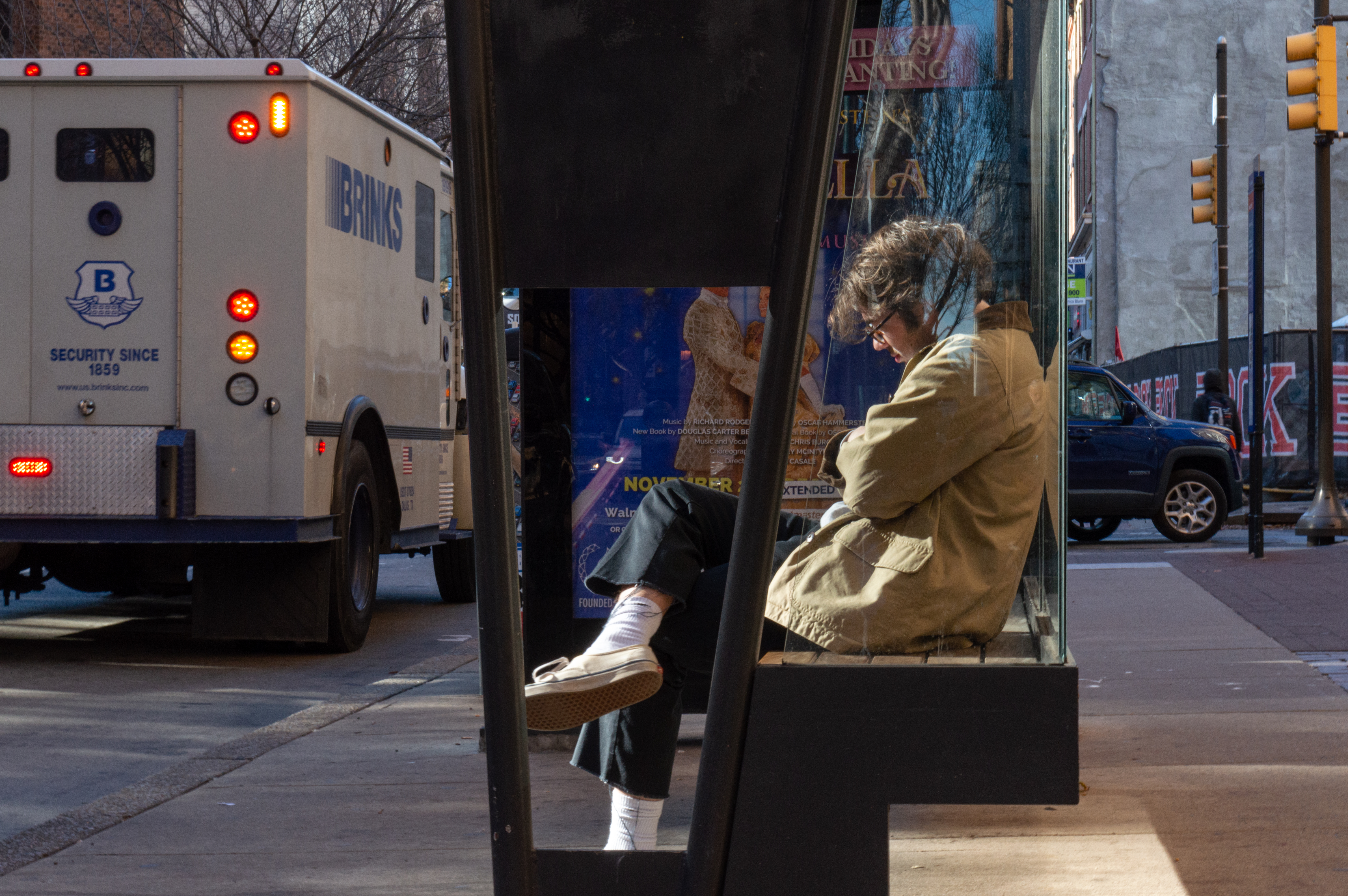 A man waits at a bus stop. This side-portrait shows him crossing his legs and arms and looking down towards the ground. On the street in front of him, a Brinks truck drives by.