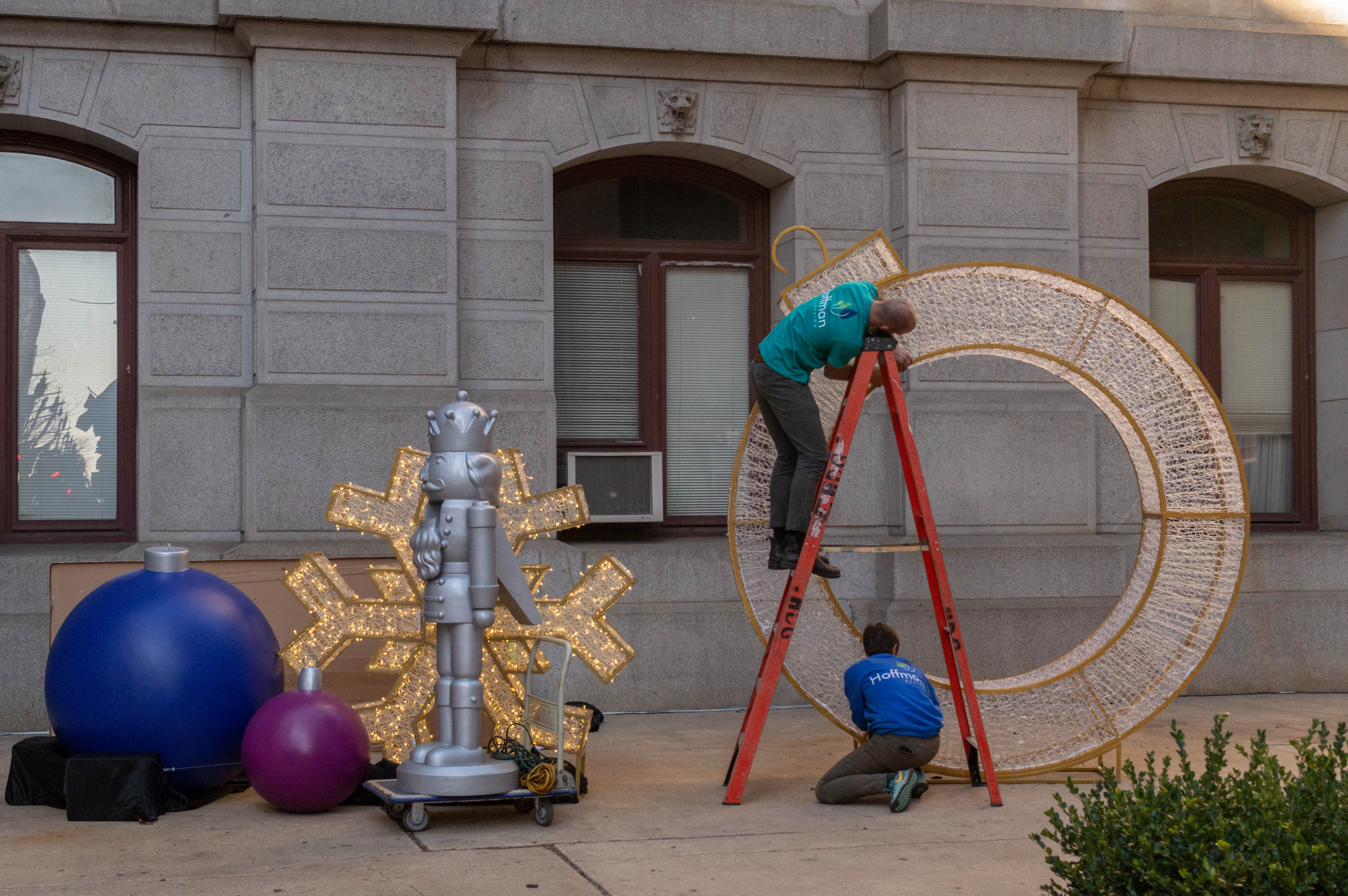 A photograph of two men setting up Christmas decorations on the sidewalk in front of a concrete building. There is a large blue Christmas tree ornament next to a large nutcracker statue in the left part of the frame. On the right, A ladder is setup, with a man crouched underneath it and a man on the top of the ladder. Both men are assembling a large circular decoration.