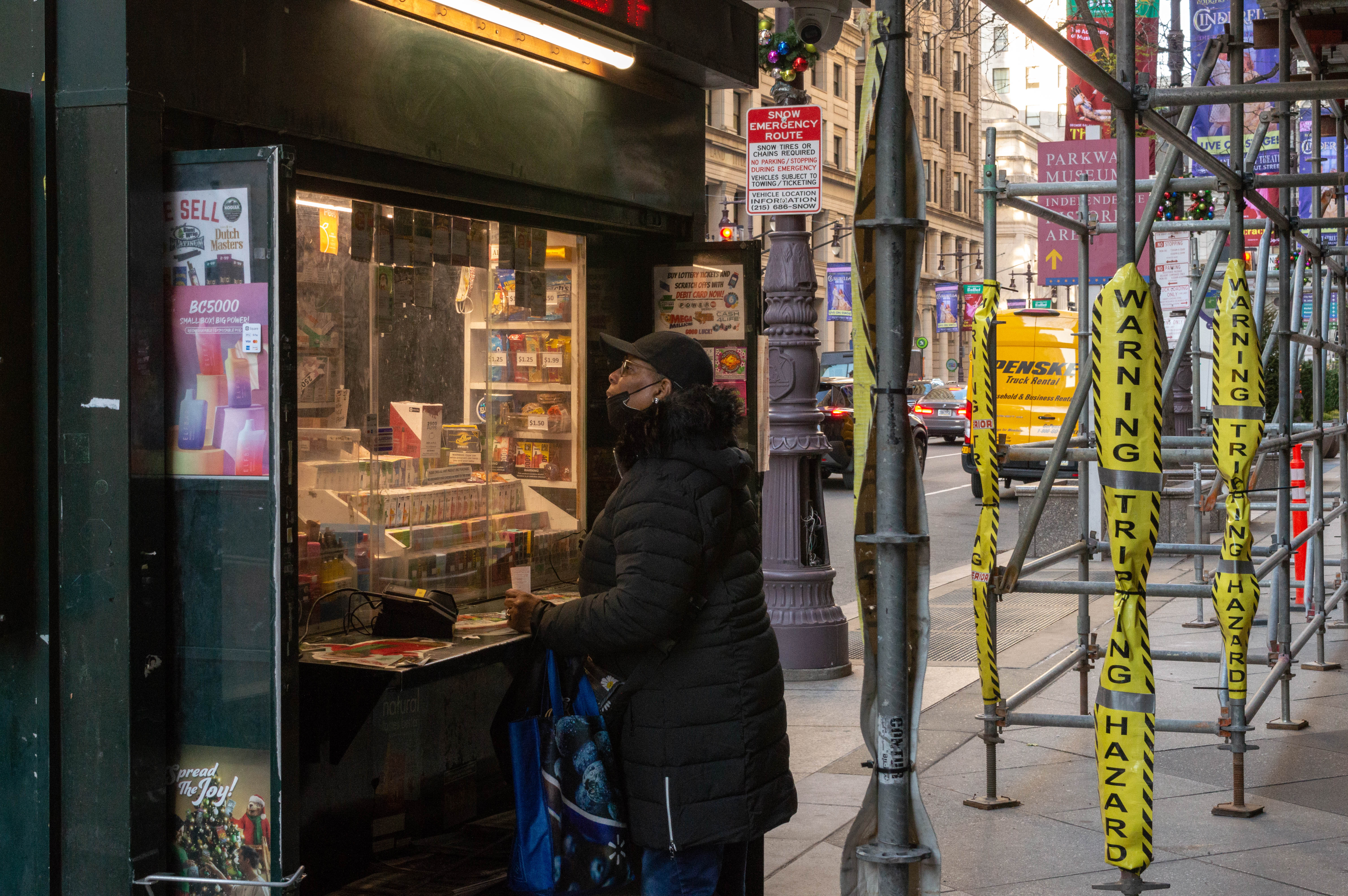 A woman stands at the counter of a newspaper stand on a city street. Surrounding the stand is scaffolding with yellow tape wrapped around its poles reading <q>Warning Tripping Hazard.<q>